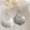 Close-up of sterling silver pear-shaped hammered dangle earrings with French hook ear wires on a marble surface.

